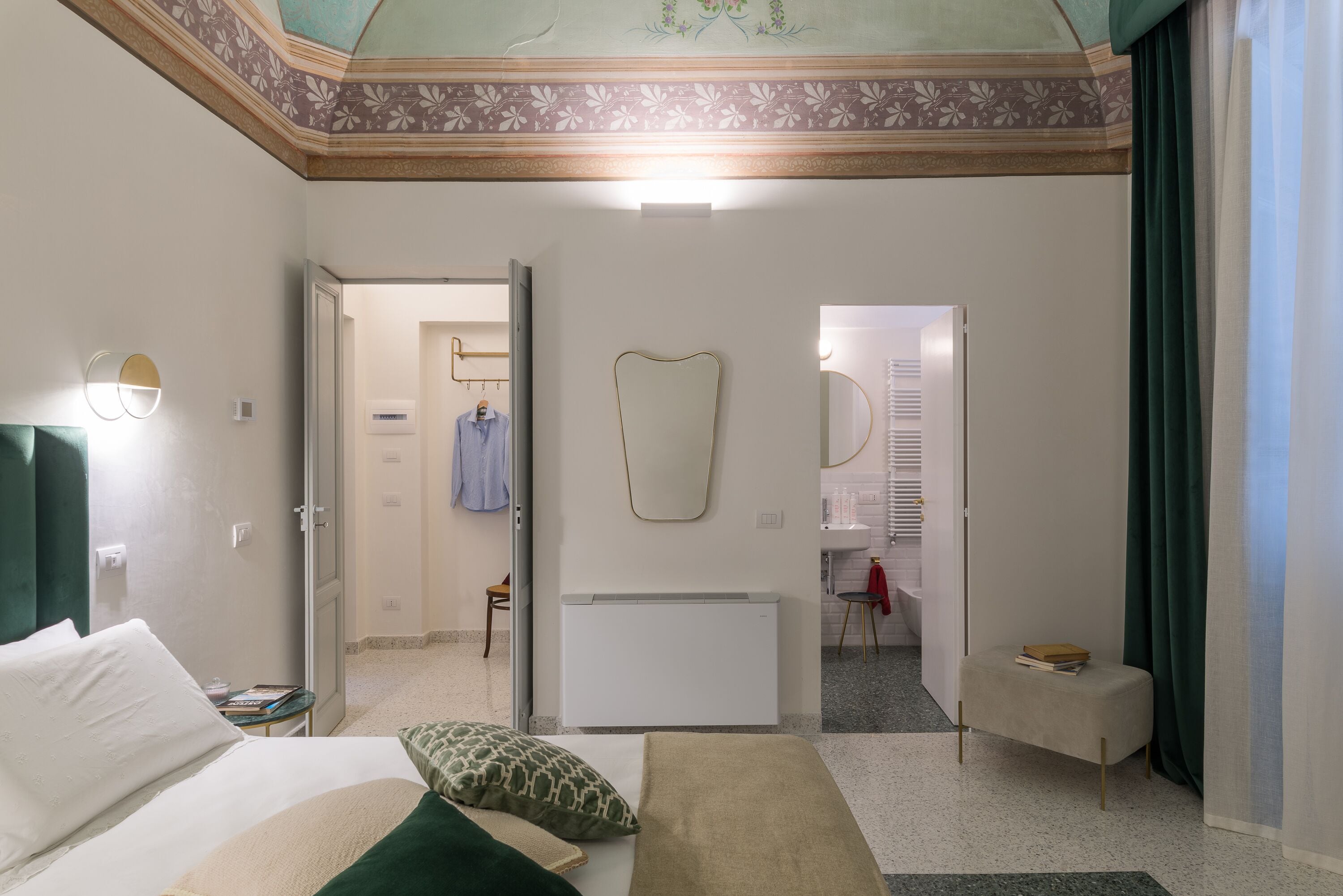 A hotel room with a bed, bedside table, a mirror, a radiator, a bench, and a door leading to a bathroom.