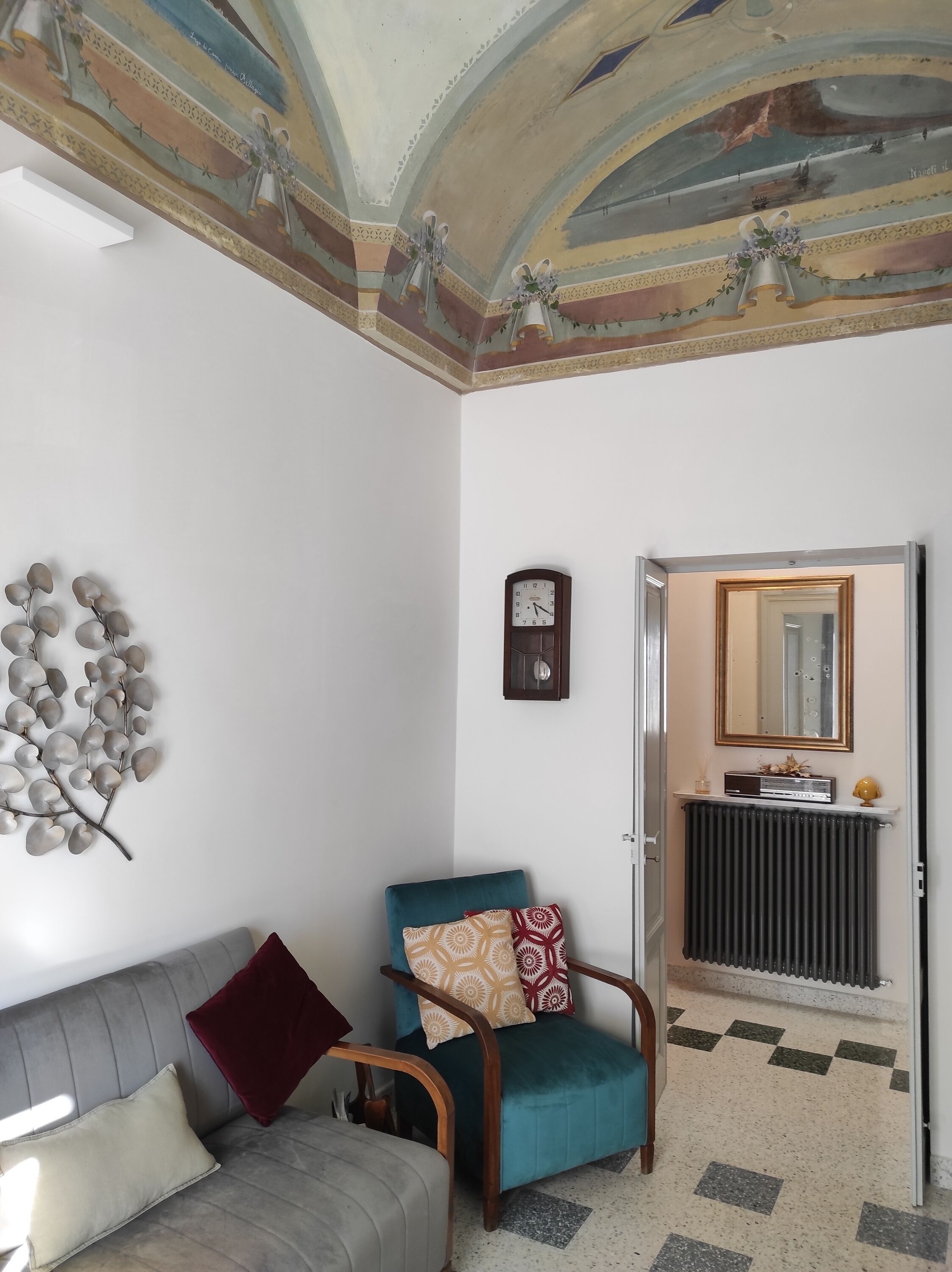 Een woonkamer met een bank, een blauwgroene fauteuil, een wandklok en een radiator.