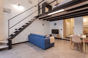 A modern living room with a staircase, a blue sofa, a dining area with a table and chairs, and a TV. - Biancospino Suite by Wonderful Italy (Noto)