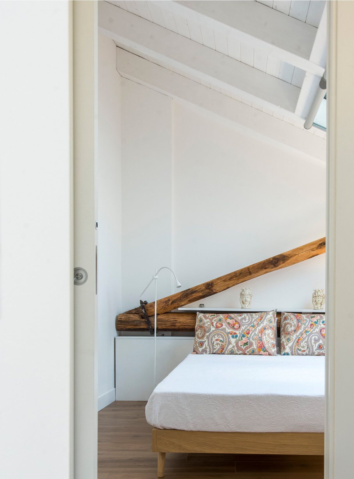 A bedroom with a bed, a wooden headboard, a white wall, a small shelf with decorative items, and a door.
