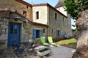 Un bâtiment en pierre avec une porte bleue, des places assises à l’extérieur et un espace jardin.