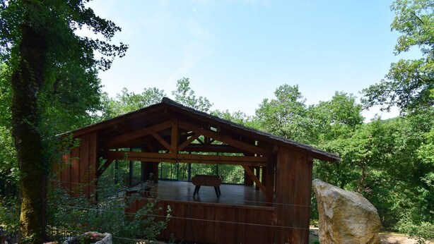 A rustic wooden cabin with a wooden deck, surrounded by dense forest.