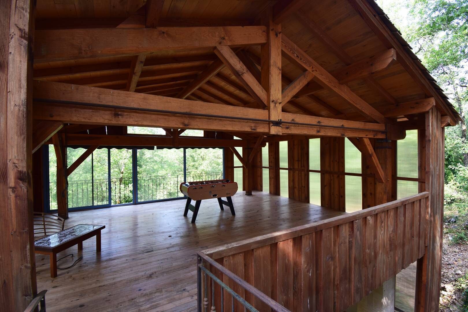 Una terraza de madera con una mesa de fútbol de mesa y un banco, rodeada de amplias ventanas.