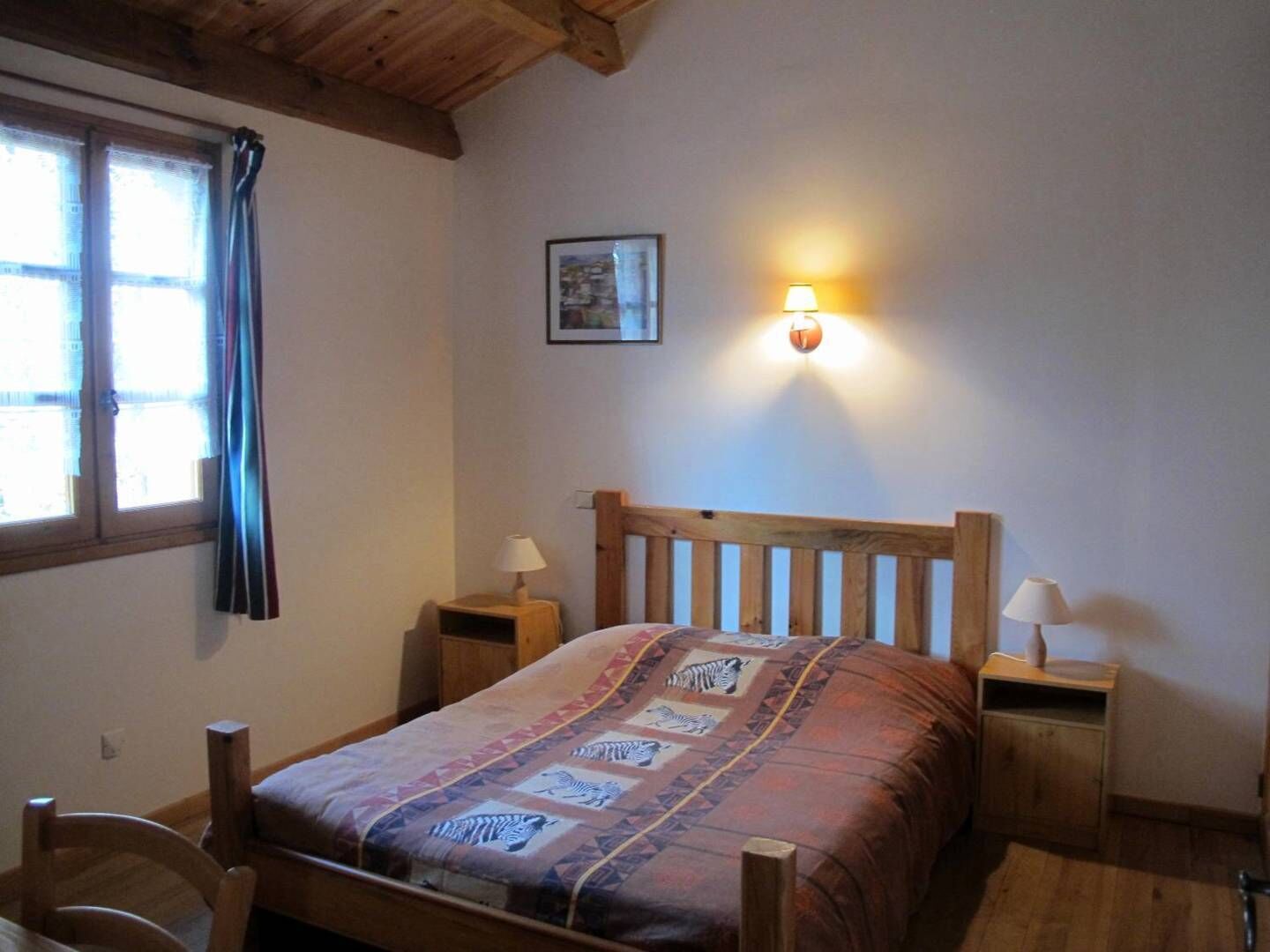 Un dormitorio con una cama de madera, dos mesitas de noche, una lámpara de pared y una ventana con cortinas, además de un cuadro enmarcado.