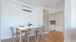 Un comedor con una mesa blanca, sillas grises y una pequeña planta en maceta. Se ve una cocina al fondo.