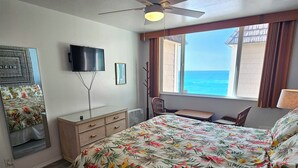 Un dormitorio con cama, televisión, ventana con vista al océano, ventilador de techo y una pequeña mesa.