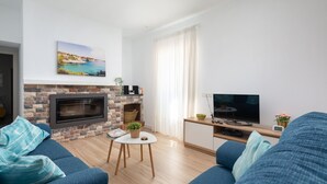 A living room with a brick fireplace, a flat-screen TV, a sofa, and a painting of a coastal scene. - CA NA MENUDA - Cosy country house with private pool in quiet surroundings. Free WiFi (Sencelles)