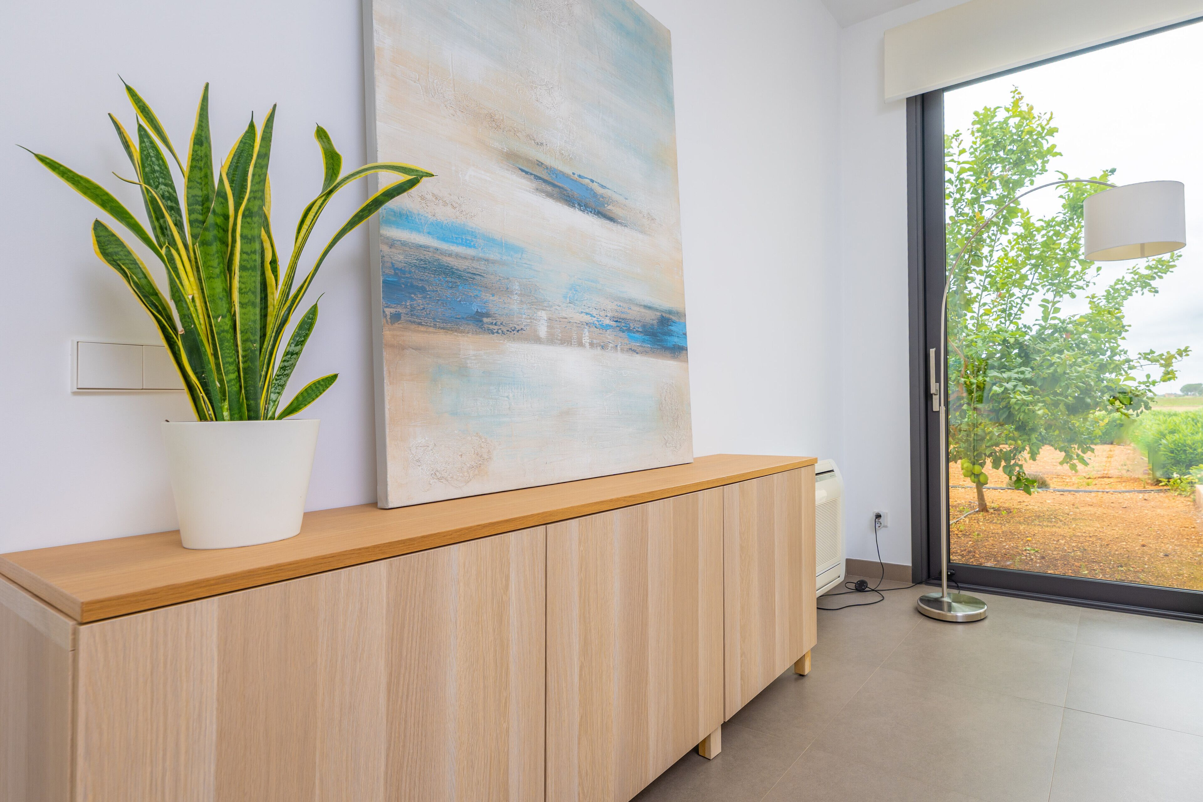 Une pièce moderne avec une console en bois, une grande peinture abstraite, une plante en pot et une vue à travers une porte vitrée.