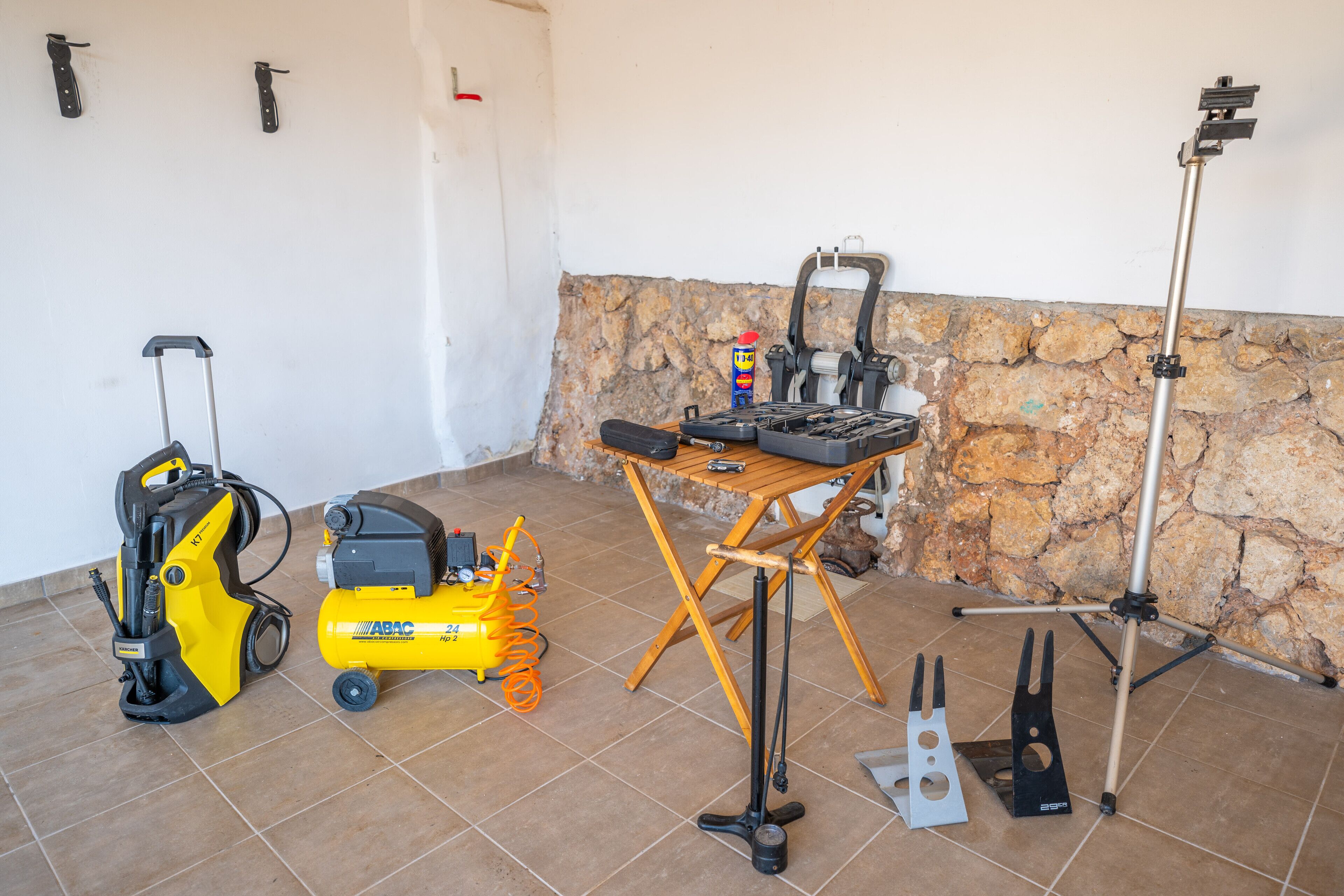 Una habitación con pared de piedra, suelo de baldosas, una mesa plegable con herramientas, una herramienta eléctrica amarilla, un aspirador y un trípode con una cámara.