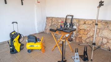 Una habitación con pared de piedra, suelo de baldosas, una mesa plegable con herramientas, una herramienta eléctrica amarilla, un aspirador y un trípode con una cámara.