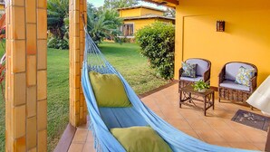 Eine Hängematte mit Kissen, zwei Korbstühle mit Sitzkissen und ein Tisch mit einer Pflanze, alles auf einer Terrasse mit gelber Wand und Blick auf einen Garten.