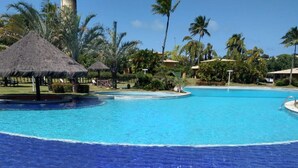 Ein Pool mit klarem, blauem Wasser, umgeben von Palmen und Reetdachschirmen.