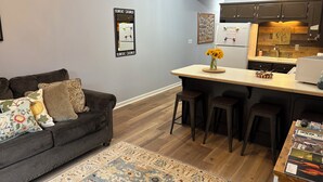 A modern living room with a dark brown sofa, a small coffee table, and a patterned rug. A kitchen area with dark wooden cabinets and a countertop is visible in the background. - Regal Ranch Retreat *DOG & HORSE FRIENDLY* (Oxford)