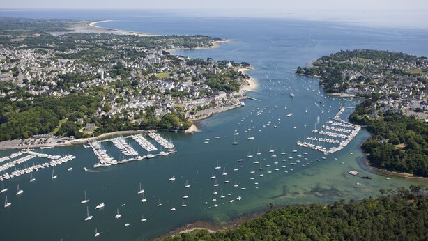 An aerial view of a coastal town with a marina filled with boats, surrounded by residential areas and a beach.
