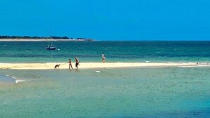 A beach with clear turquoise water, a sandy shore, and a sailboat in the distance.