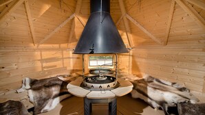 A wooden cabin interior with a central wood stove surrounded by fur-covered benches.