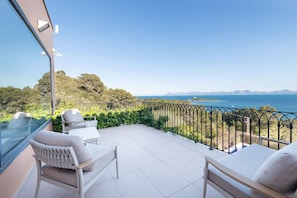 A balcony with white chairs, a glass wall, and a view of the sea and mountains.