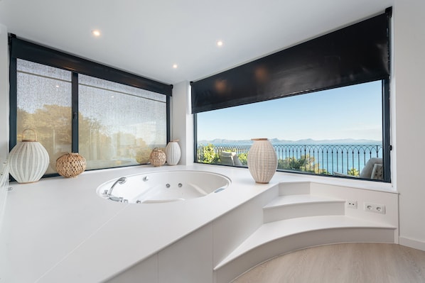 A modern bathroom with a large bathtub, a window with a view of the sea, and decorative vases on the counter.