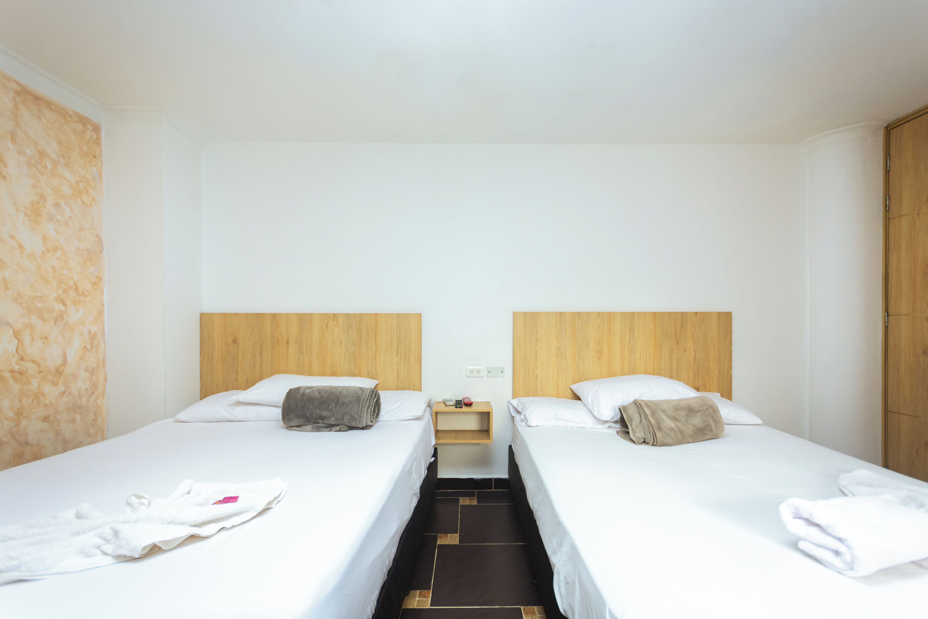A hotel room with two single beds, a wooden headboard, a nightstand with a lamp, and a tiled floor.