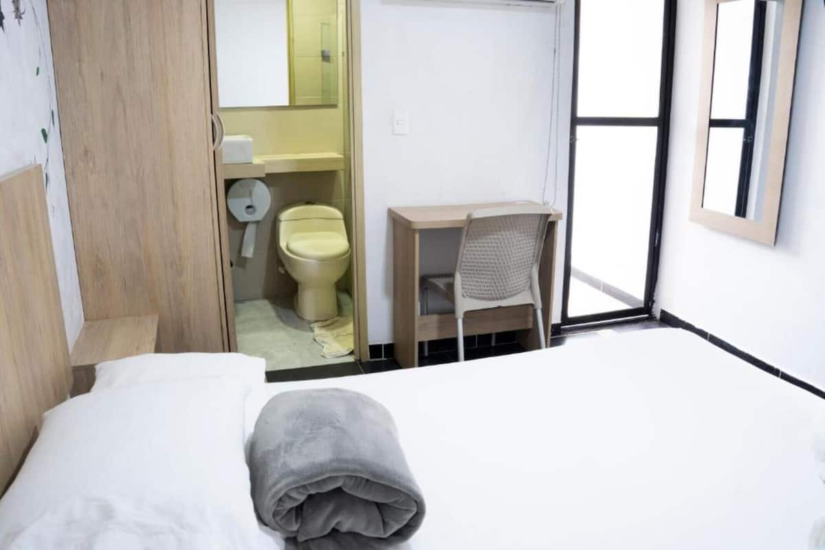 A hotel room with a bed, a desk, a chair, a toilet, and a window.