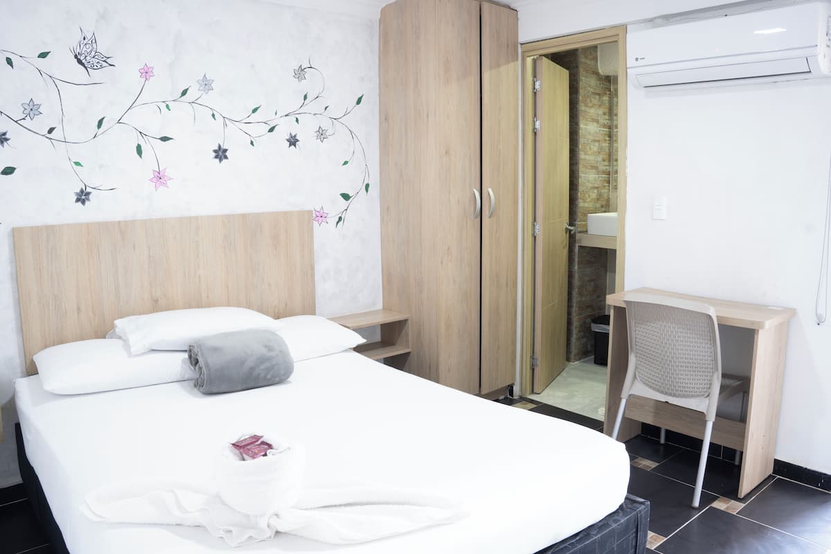 A hotel room with a bed, a wooden wardrobe, a desk with a chair, and a bathroom visible through a glass door.