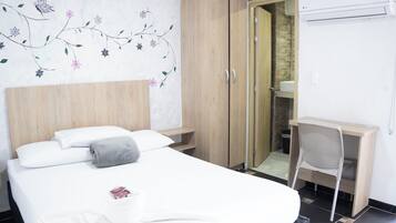 A hotel room with a bed, a wooden wardrobe, a desk with a chair, and a bathroom visible through a glass door.