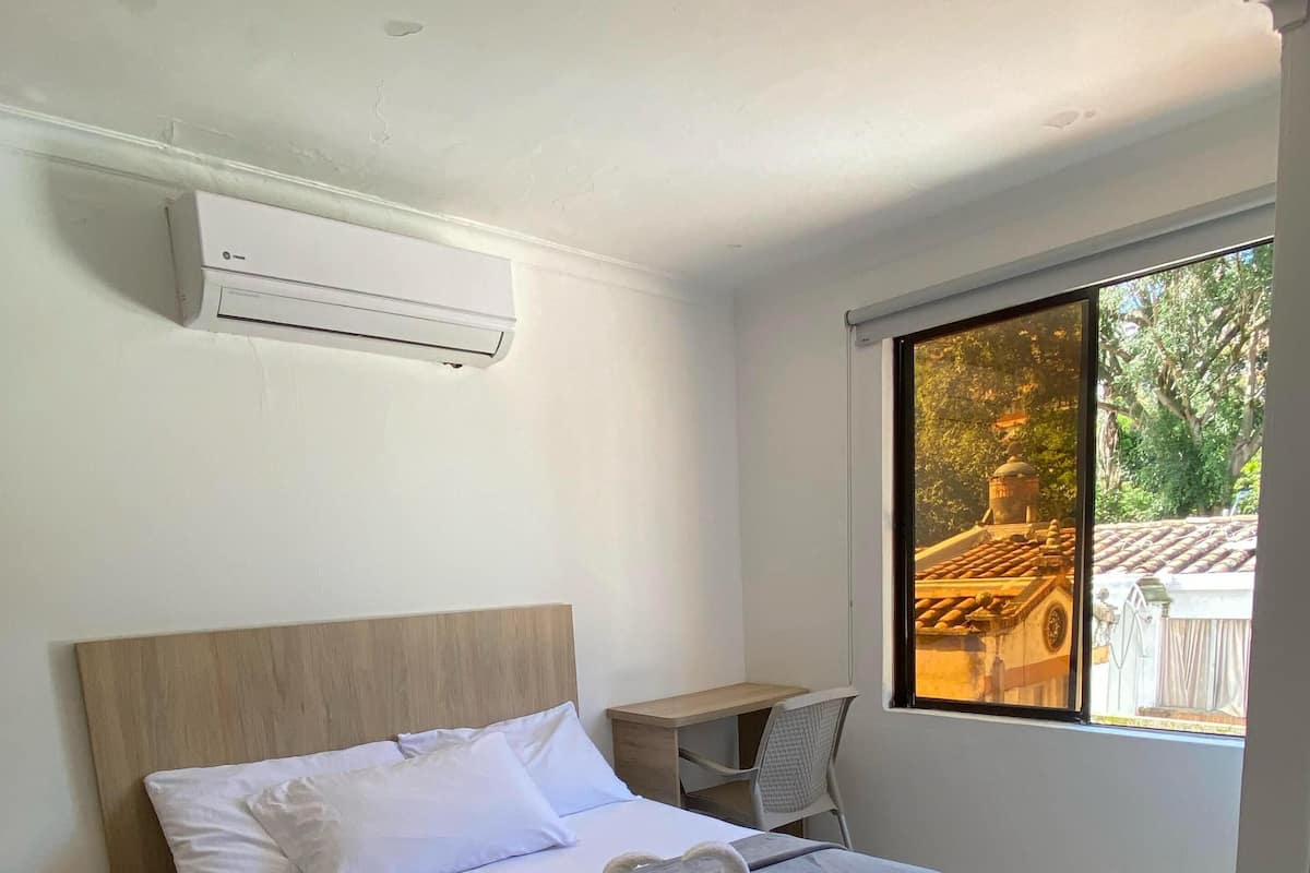 A hotel room with a bed, a desk, and a window with a view of a building and trees.
