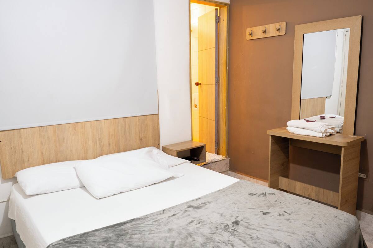 A hotel room with a bed, a wooden headboard, a bedside table, a nightstand, a mirror, and a wooden shelf with towels.