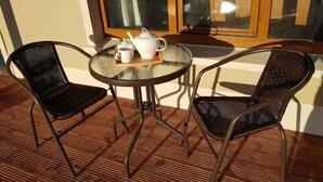 Una mesa pequeña y redonda con dos sillas de mimbre, una bandeja con tetera y tazas, y una ventana con cortinas.