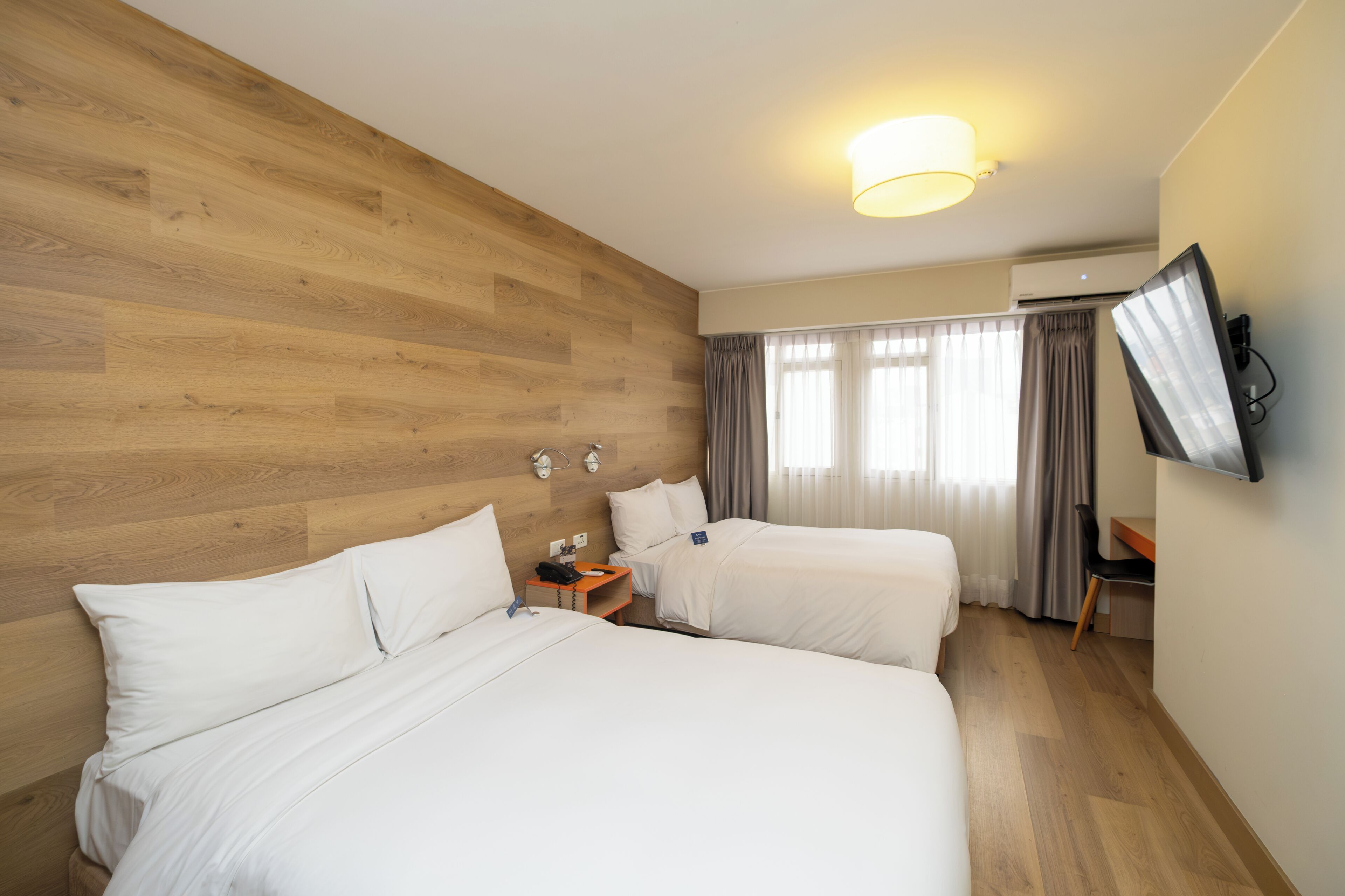 Habitación de hotel con dos camas, un escritorio, una silla, un televisor en la pared y una ventana con cortinas.