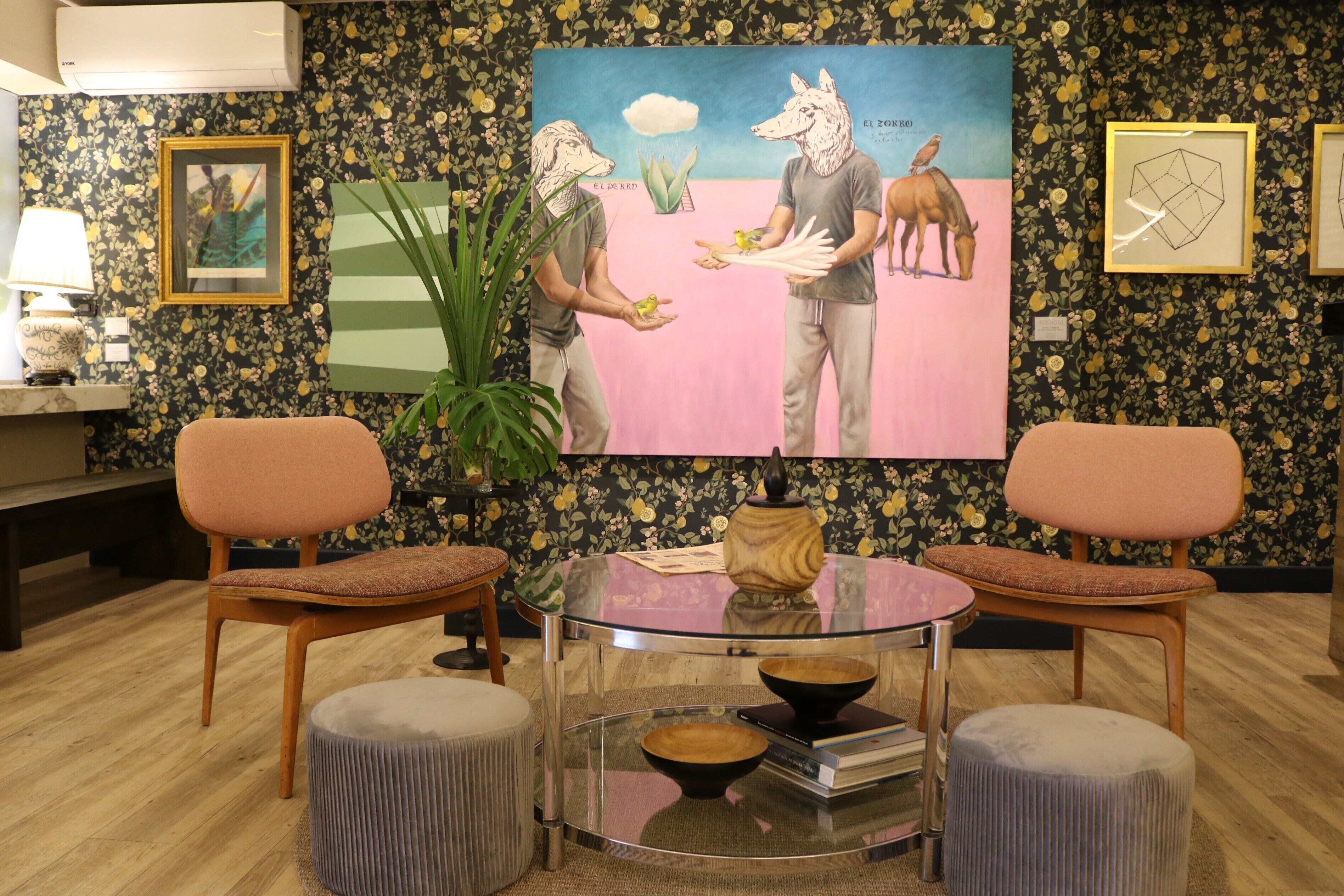 A room with two wooden chairs, a glass table with a bowl and books, a framed picture on the wall, and a floral wallpaper pattern.