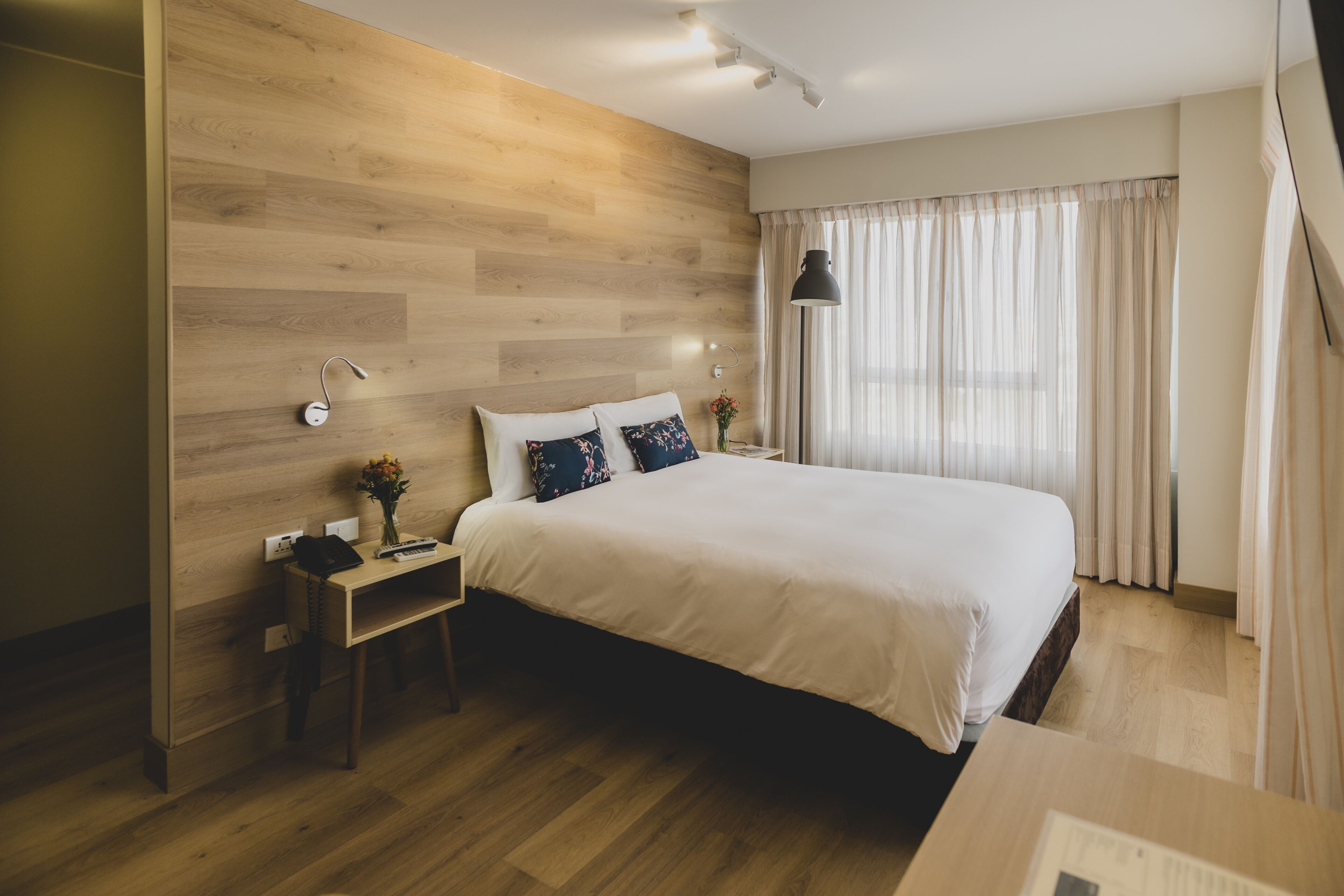 Moderni hotellihuone, jossa on puukoristeinen seinä, suuri sänky valkoisilla vuodevaatteilla, yöpöytä lampun kanssa ja näkymä ulos ohuiden verhojen läpi.