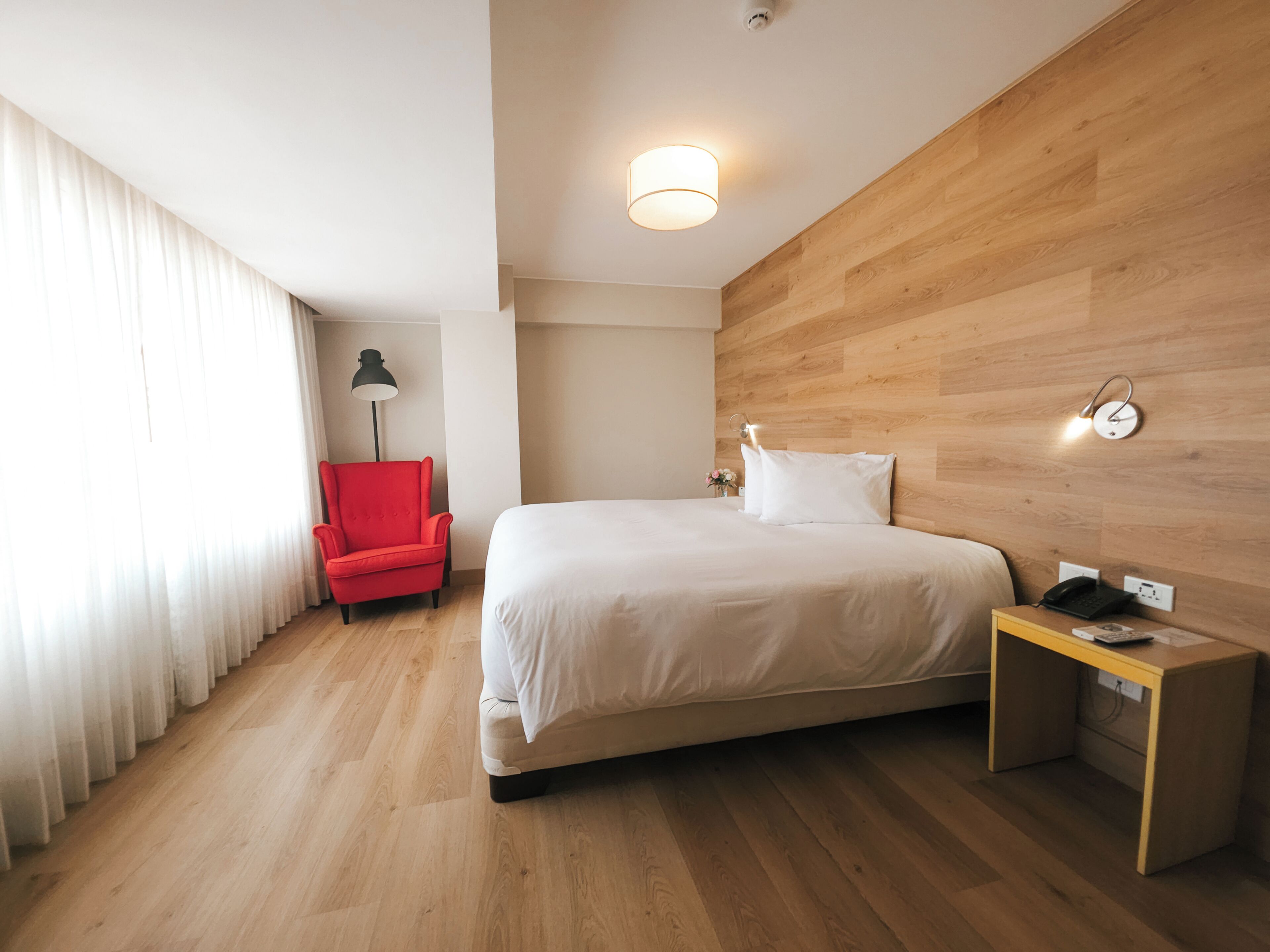 A hotel room with a wooden floor, a bed with white linens, a red armchair, a nightstand with a phone, and a wall-mounted light fixture.