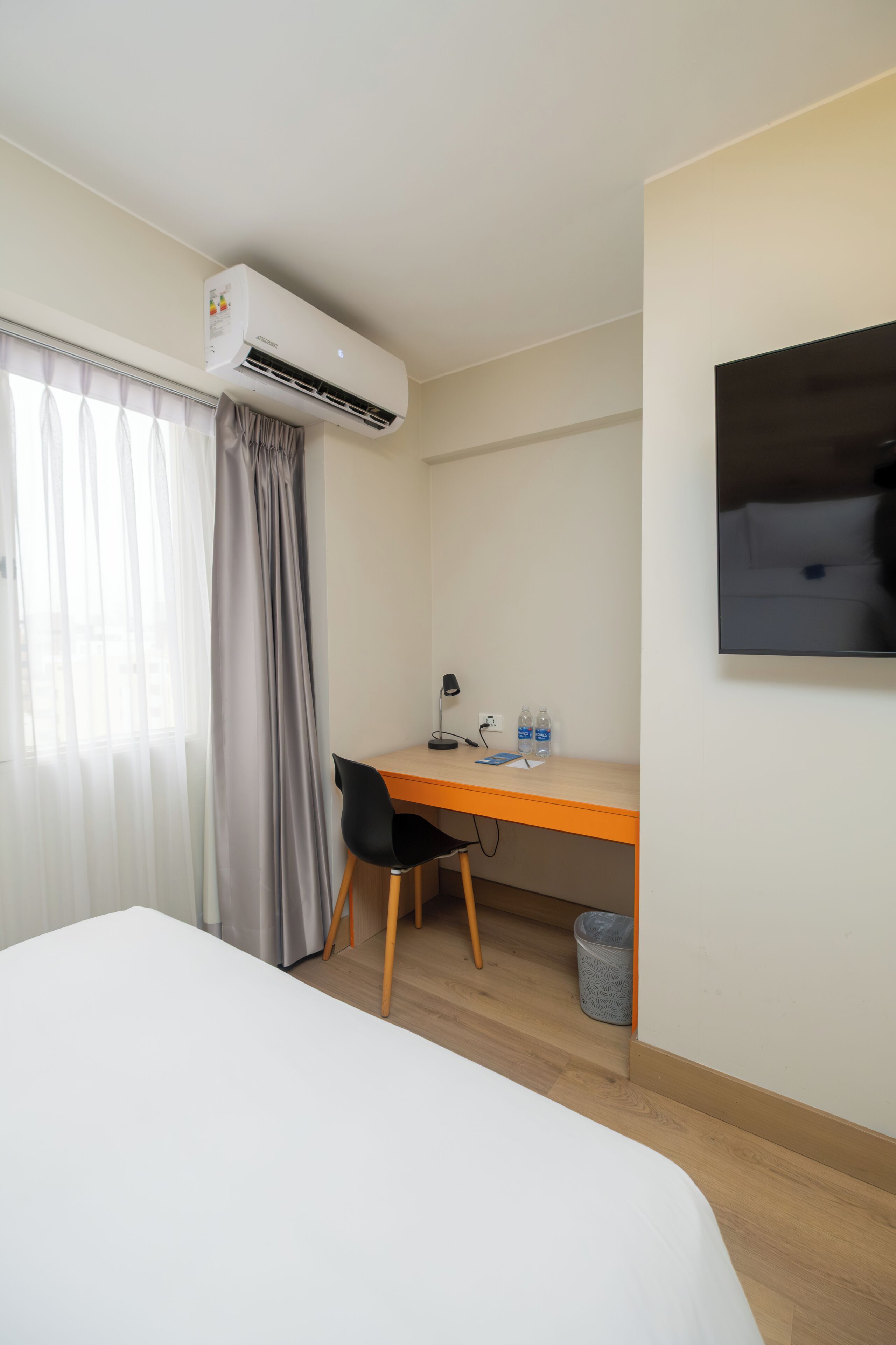 Habitación de hotel con cama, escritorio con silla, aire acondicionado, televisor y ventana con cortinas.