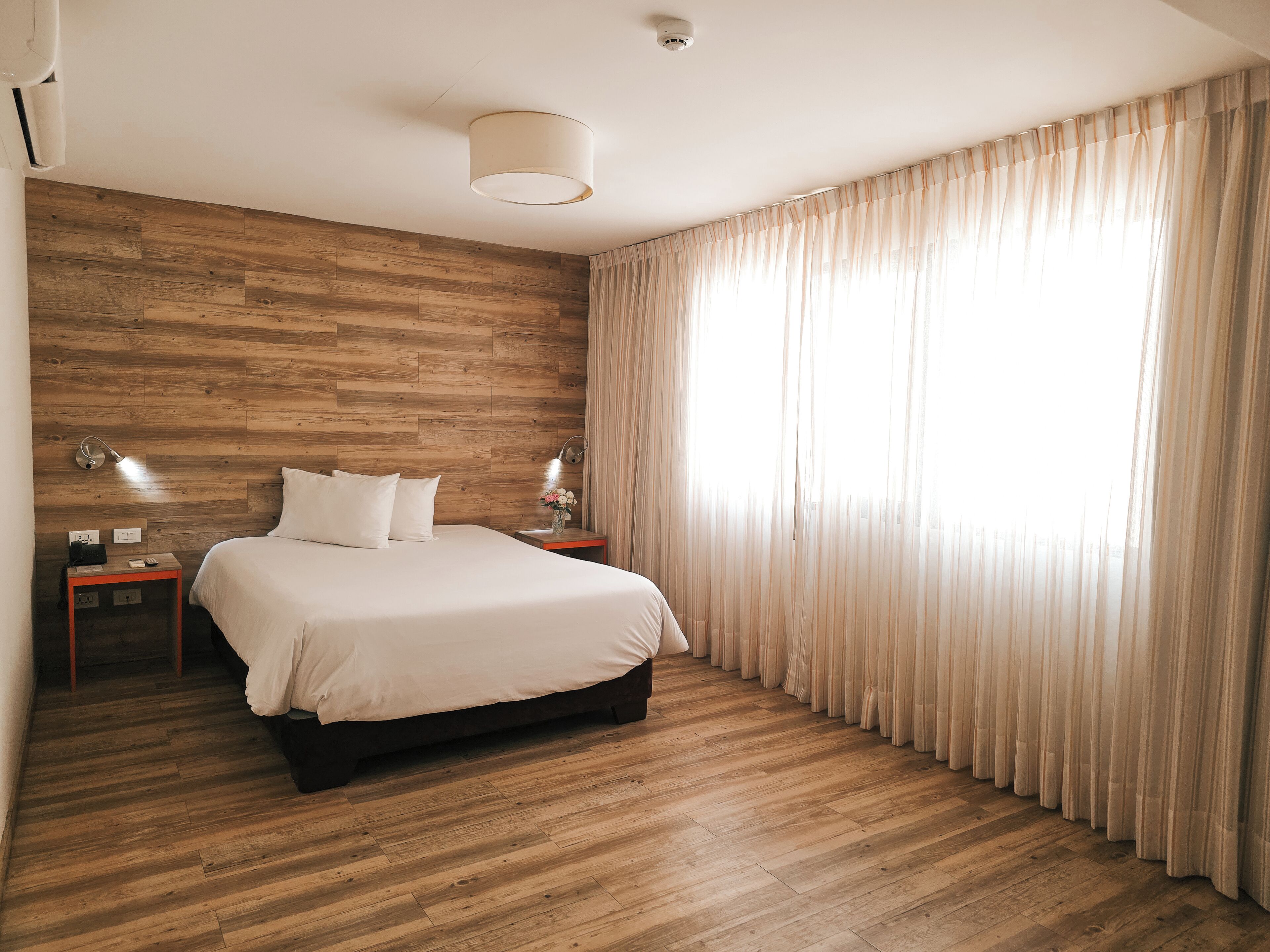 Habitació d'hotel amb terra de fusta, un llit amb roba de llit blanca, dues taules de nit amb làmpades i cortines translúcides a la finestra.