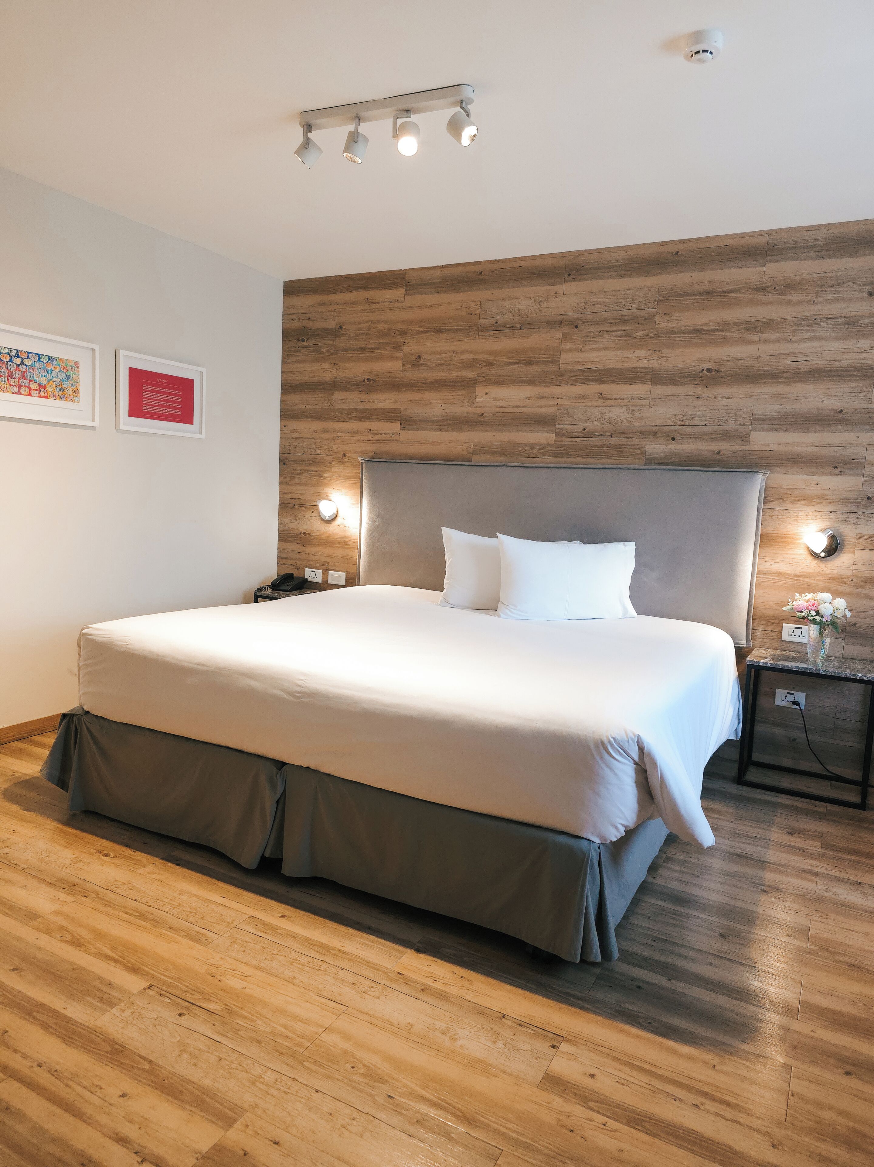 Un lit bien fait, avec une tête de lit grise, un plancher en bois et un luminaire fixé au mur au-dessus.