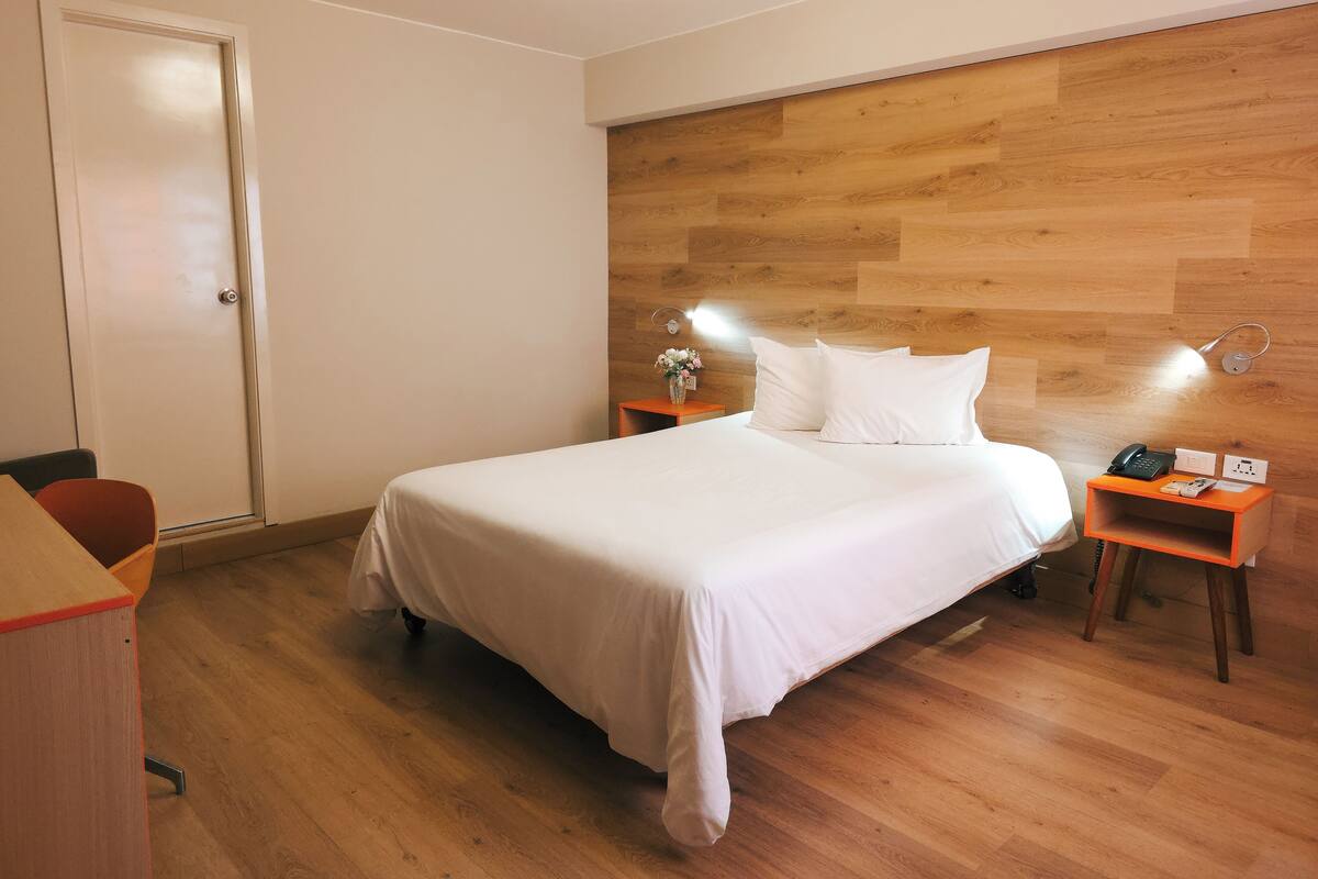 Una habitación de hotel con tablero de cabecera de madera, cama con ropa blanca, mesita de noche con lámpara y escritorio con silla.