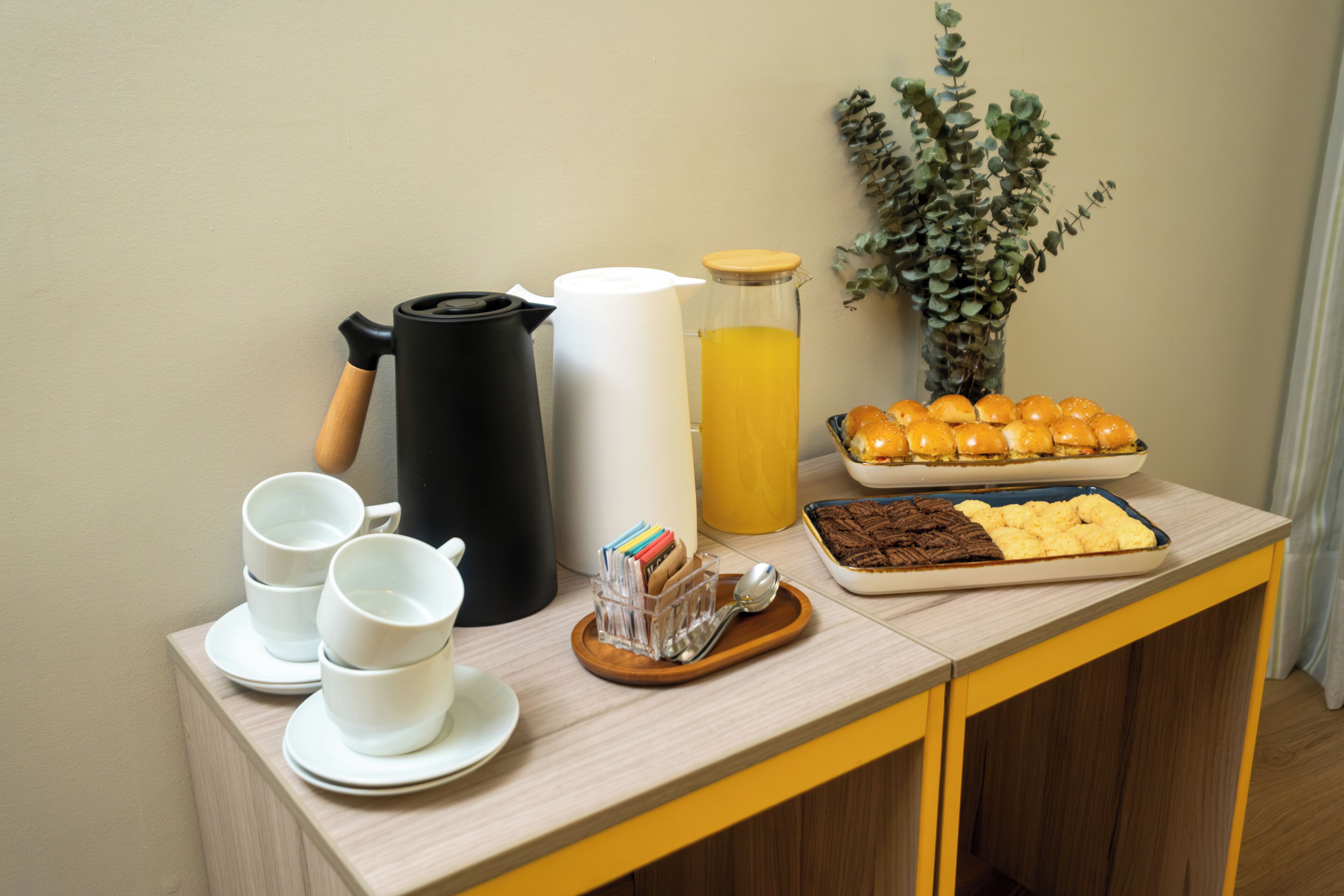 Uma mesa de madeira com máquina de café, sumo, pastelaria e chávenas.