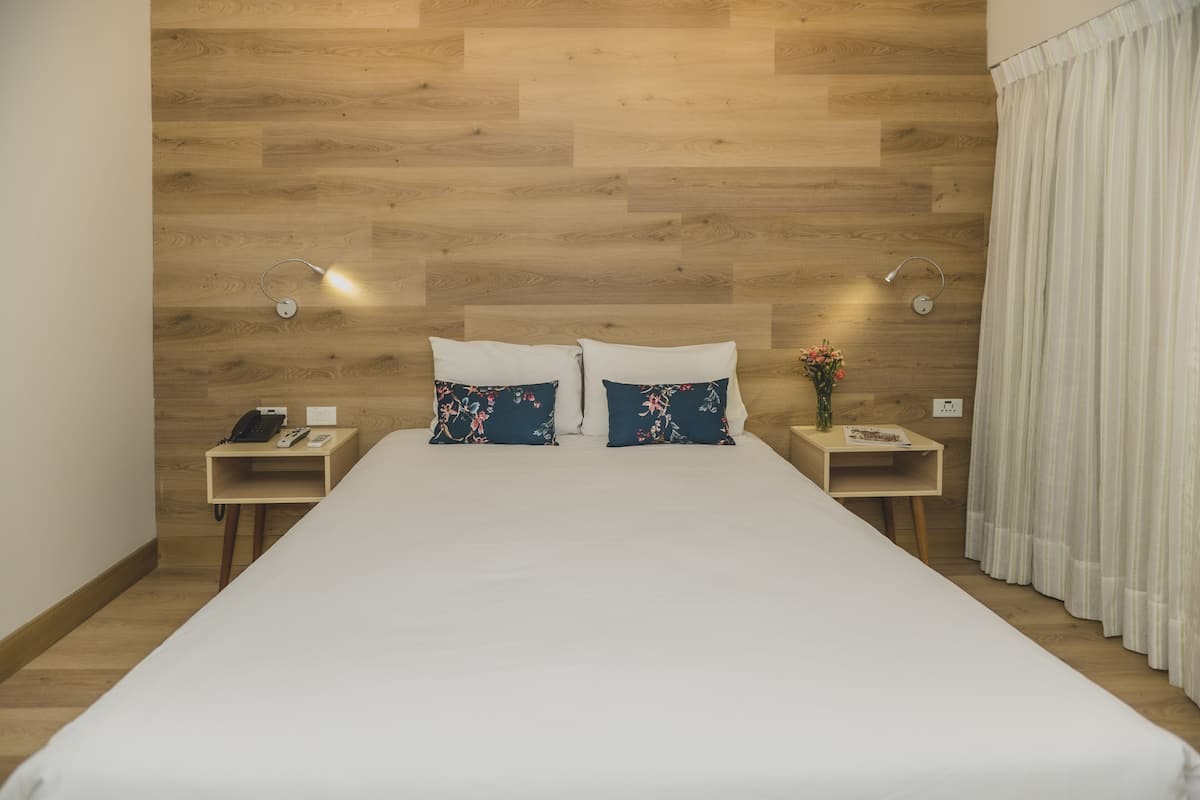 Habitación de hotel con pared de madera, una cama con ropa blanca, dos mesitas de noche con lámparas y un ventanal con cortinas.