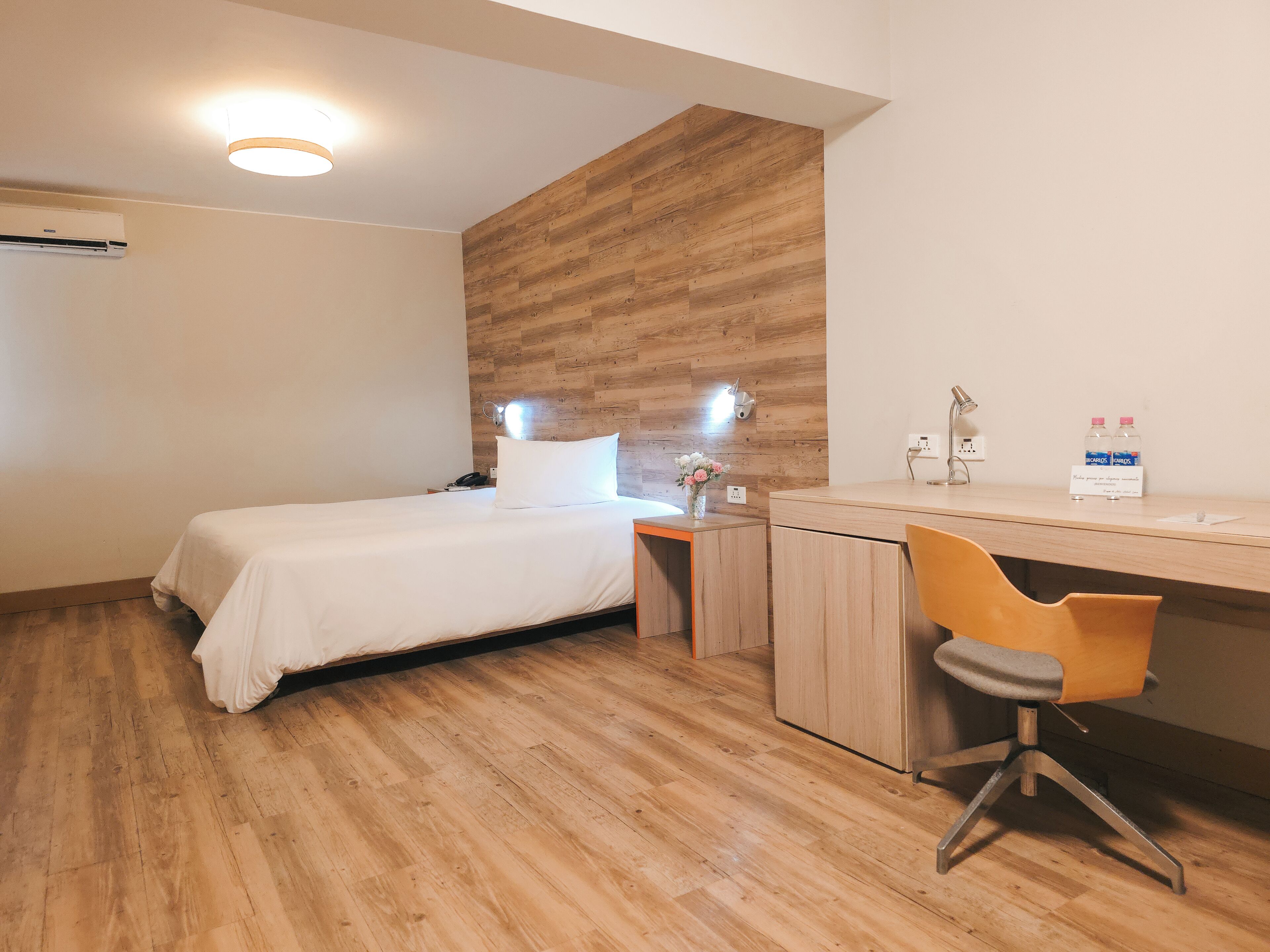 A hotel room with a bed, a desk, a chair, a nightstand, a wall-mounted lamp, and a water bottle on the desk.