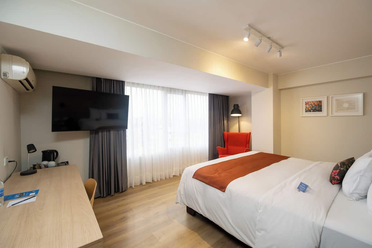 Habitación de hotel con cama, escritorio, silla, televisor, lámpara y ventana con cortinas.