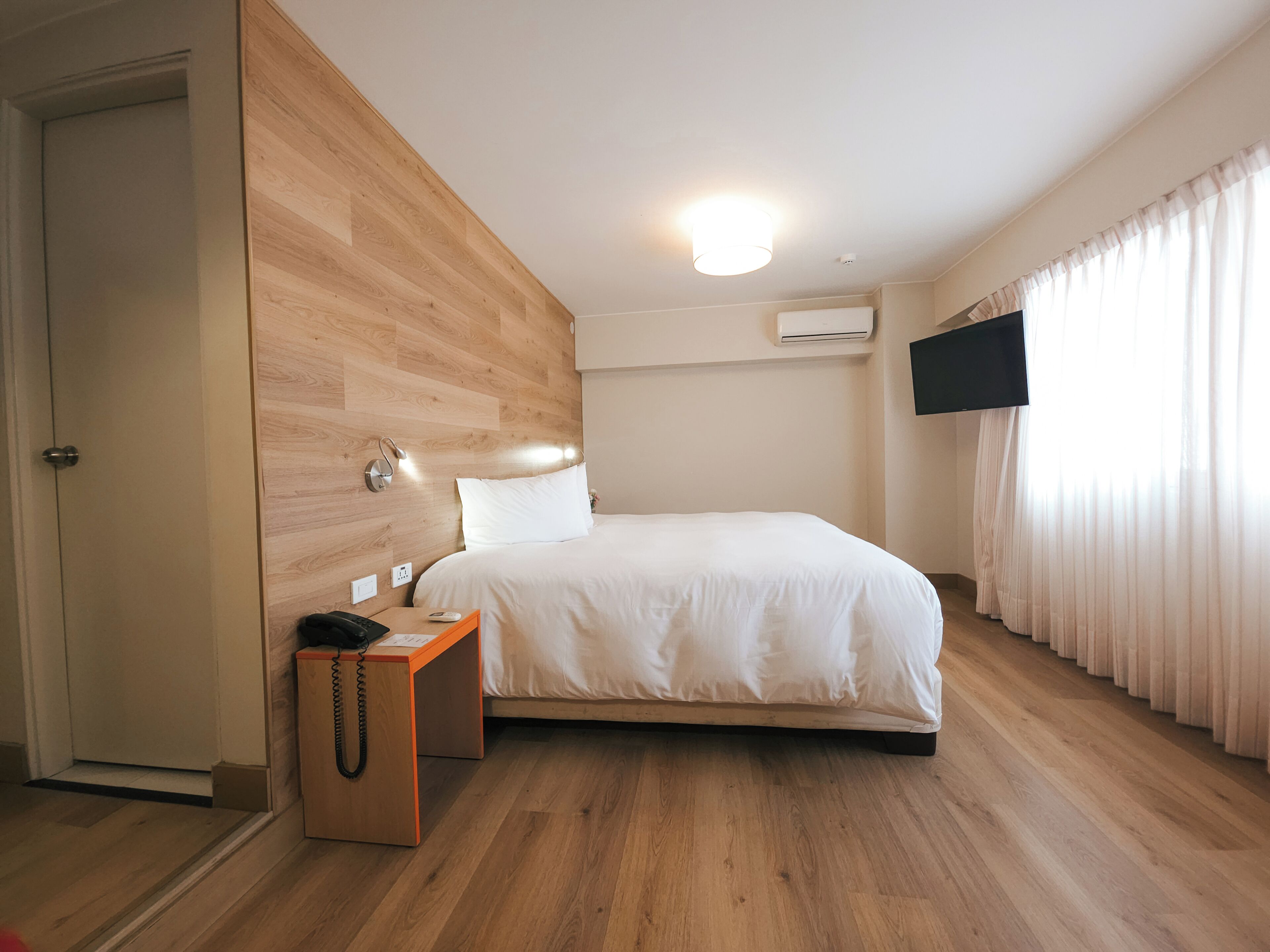 A modern hotel room with a wooden accent wall, a single bed, a bedside table with a telephone, and a flat-screen TV mounted on the wall.