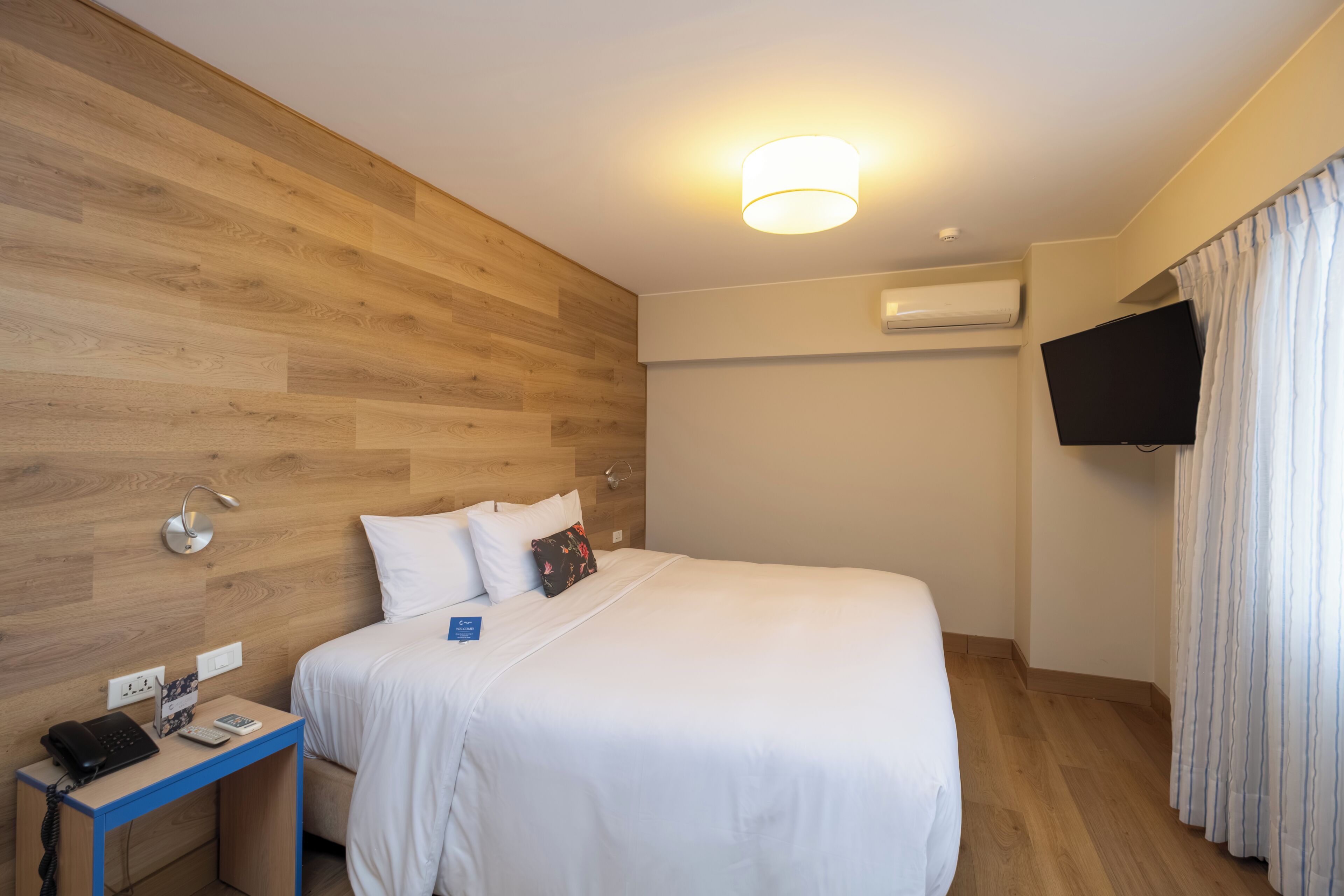 Hotelkamer met een houten wand, een bed met wit beddengoed, een nachtkastje met telefoon en lamp, een airconditioner en een aan de muur bevestigde televisie.