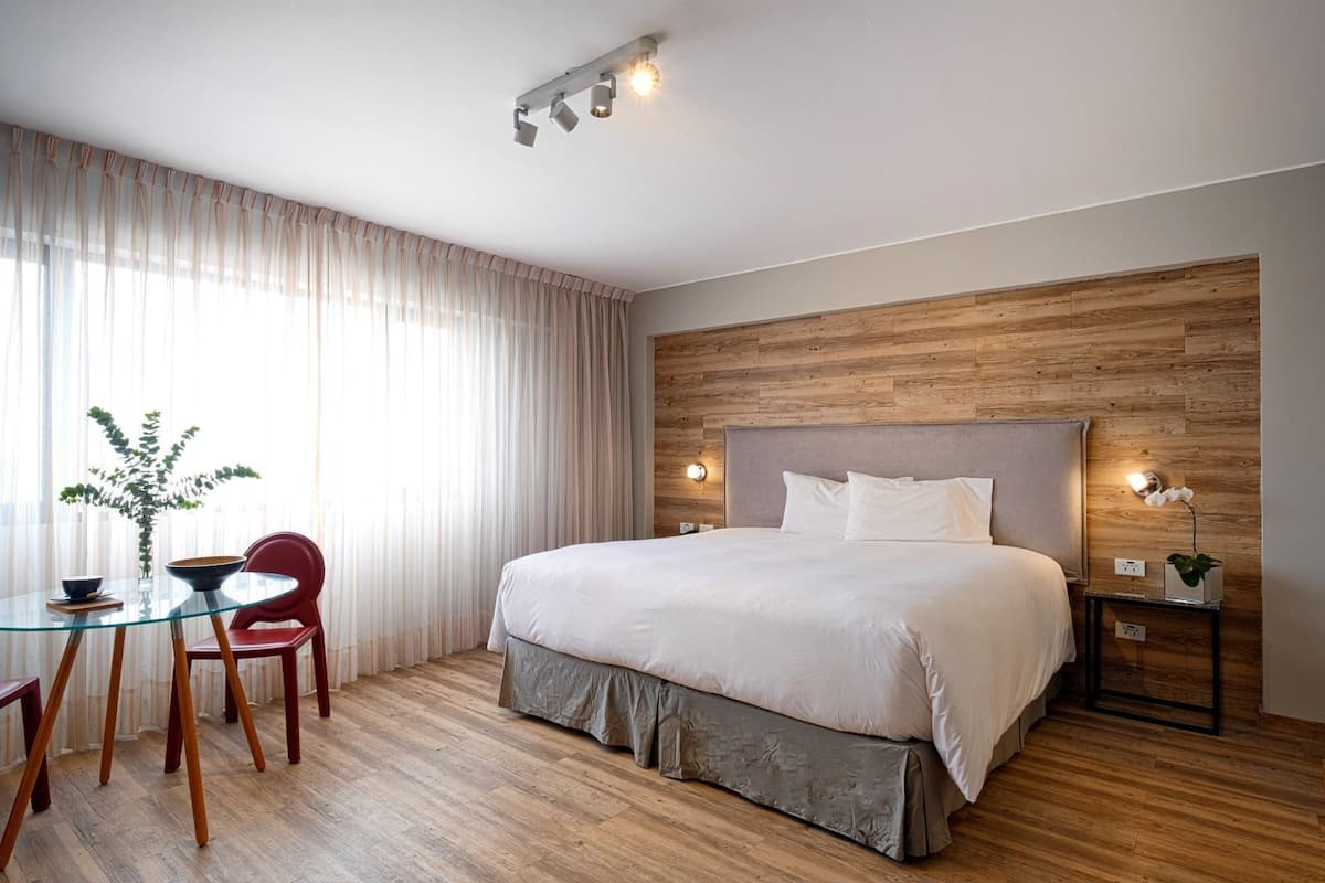 Una habitación de hotel con una cama, una mesita de vidrio con sillas, una planta en maceta y una pared de madera.