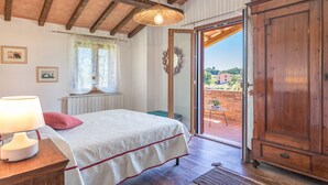A bedroom with a bed, a bedside table with a lamp, a window with curtains, and a view of a courtyard through an open door.