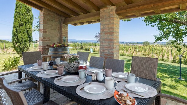 An outdoor dining area with a table set for four, surrounded by wicker chairs, under a wooden pergola with a view of vineyards and hills.