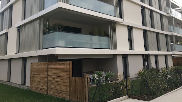 A modern multi-story residential building with balconies, a wooden gate, and a well-maintained garden.