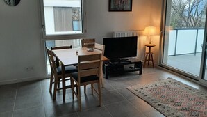 A living room with a TV, a dining table with chairs, a lamp, a world map clock, and a large window.