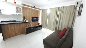A modern living room with a sofa, a TV, a wooden cabinet, and a kitchen area. - 229, Apartamento 2 Dormitórios em Local Agradável (SC)