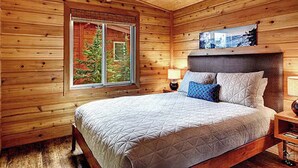 A wooden cabin room with a bed, a window with a view of trees, and a mounted picture on the wall.